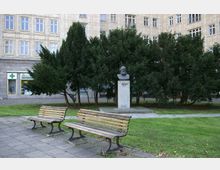 Büste von Karl Marx auf einem steinernen Sockel, umgeben von hohen Bäumen in einem Platz mit Grünflächen und Pflastersteinen; im Hintergrund ein mehrstöckiges Gebäude mit regelmäßig angeordneten Fenstern und Geschäften im Erdgeschoss.