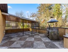 Überdachte Terrasse mit gemustertem Steinboden, Holzdecke und Metallstützen, umgeben von niedrigen Wänden und Gitterelementen. Rechts steht ein gemauerter Grill- oder Kaminaufbau mit Metallhaube, dahinter Bäume und ein Zaun im Außenbereich.