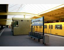 Eine U-Bahn-Station mit modernem Glasdach, hellgelber Metallkonstruktion und einer grün gekachelten Wand. Im Vordergrund steht ein Stationsschild mit der Aufschrift „Onkel Toms Hütte“ und Sitzbänken, während eine gelbe U-Bahn am Bahnsteig hält.