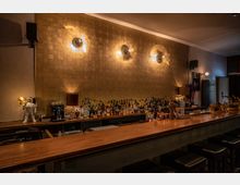 Eine stilvolle Bar mit langer Holztresenfläche und gepolsterten Hockern, dahinter ein Regal mit einer großen Auswahl an Spirituosen. Die Wand ist mit schimmernden, goldfarbenen Kacheln gestaltet, beleuchtet von modernen Wandleuchten, die kreisförmige Lichtmuster werfen.