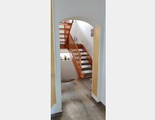 Eine innenliegende Treppe aus hellem Holz mit offenen Stufen führt in eine obere Etage. Der Raum hat hell gestrichene Wände, einen Boden in Holzoptik und gelbe Wandakzente. Ein Rundbogen rahmt die Sicht auf die Treppe.
