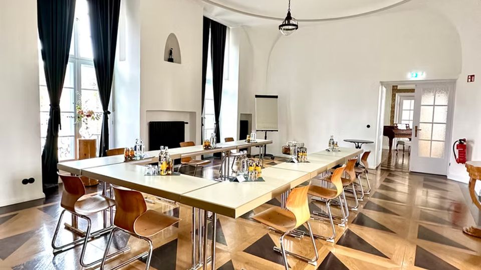 Heller Konferenzraum mit U-förmig angeordneten Tischen und Holzstühlen auf einem geometrisch gemusterten Parkettboden. Hohe Fenster mit dunklen Vorhängen und eine auffällige, spiralförmige Stuckdecke mit Hängelampe prägen den Raum; seitlich befindet sich eine Tür zu einem angrenzenden Bereich.