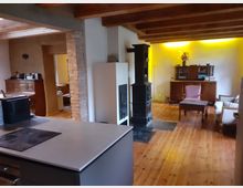 Wohnzimmer in einem rustikalen Stil mit Holzboden, freiliegenden Holzbalken und einem schwarzen Kaminofen in der Mitte. Im Hintergrund stehen Holzmöbel, darunter ein Schrank und ein Stuhl, sowie eine indirekte gelbe Beleuchtung an der Wand.