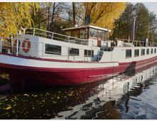 Ein langes weißes und rotes Schiff liegt an einem Ufer, umgeben von herbstlich gefärbten Bäumen. Die Wasseroberfläche reflektiert das Schiff und die umliegende Landschaft.