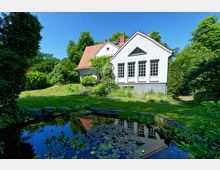 Weißes Gebäude mit Satteldach in einem üppig begrünten Garten, umgeben von Bäumen. Im Vordergrund ein kleiner Teich mit Seerosen und Steinrandung.