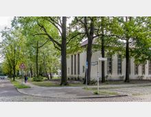 Straßenecke mit einem Fußweg, gesäumt von hohen Bäumen und einem Gebäude mit großen, schmalen Fenstern; Straßenschilder „Isoldestraße“ und „Dreilindenstraße“ sowie ein Verkehrsschild „Achtung Kinder“ sind sichtbar. Im Hintergrund befinden sich weitere Bäume und parkende Autos.