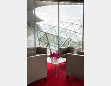 Sitzbereich mit vier grauen Sesseln um einen kleinen, weißen Tisch, auf dem rote Blumen in Glasvasen stehen. Im Hintergrund große Fenster mit Blick auf eine moderne Glaskuppel-Konstruktion und den Himmel.