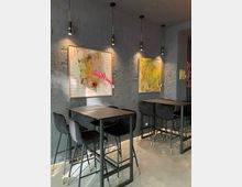 Ein modernes Café mit schwarzen Hochstühlen und Tischen auf einem grauen Betonboden. Die Wände sind dunkelgrau und mit abstrakten, farbenfrohen Bildern dekoriert; von der Decke hängen schlichte schwarze Hängeleuchten.