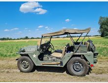 Ein olivgrüner US-Armee-Geländewagen mit offener Kabine steht auf einem Feldweg vor einer weiten Wiesen- und Feldlandschaft. Im Hintergrund sind Heuballen, Bäume und ein klarer, blauer Himmel mit vereinzelten Wolken zu sehen.