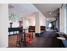 Ein moderner Innenraum mit Holzfußboden, hohen Decken und schlichter Beleuchtung. Der Raum enthält Stehtische mit Hockern, eine Sitzbank mit roten Polstern an der Wand, und dekorative Elemente wie eine grüne Pflanzenmuster-Tapete und Fenster, die Tageslicht hereinlassen.