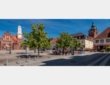 Großer gepflasterter Marktplatz mit mehreren Laubbäumen, Sitzbänken und Fahrradständern, umgeben von historischen Gebäuden mit roten Ziegeldächern. Im Hintergrund ragen ein Backstein-Rathaus mit hellem Uhrturm sowie ein weiterer Turm mit grüner Haube über die Platzbebauung.