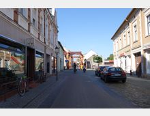 Schmale gepflasterte Straße in einer Altstadt mit historischen Gebäuden auf beiden Seiten, darunter ein Restaurant mit Schaufenstern links. Im Hintergrund ist eine Metallkonstruktion sowie eine Treppe zu sehen, während Fahrräder geparkt und Autos am Straßenrand stehen.