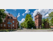 Backsteingebäude mit einem Turm in historischem Stil, umgeben von Bäumen und einer gepflasterten Straße. Im Vordergrund ein Straßenschild mit der Aufschrift „Parksiedlung“, im Hintergrund parkende Autos und weitere Backsteingebäude.