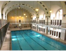 Hallenbad mit einem großen rechteckigen Schwimmbecken in der Mitte, umgeben von Kacheln und Umkleideräumen. Der Raum hat hohe, gewölbte Decken mit hängenden Lampen und großen Rundbogenfenstern, die Tageslicht hereinlassen.