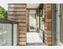 Moderne Balkonfläche mit Holzverkleidung an den Wänden, großen Glas-Schiebeelementen und einem Geländer aus Glas und Metall. Im Hintergrund stehen Pflanzkübel mit grünen Pflanzen auf einem hellen, gefliesten Boden.