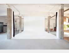 Minimalistisches Fotostudio mit einem großen, weißen Hintergrund, umgeben von sichtbaren Betonstützen. Der Raum ist offen gestaltet, mit einfachem Mobiliar und einem Tischbereich im Hintergrund.