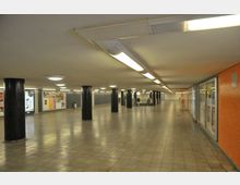 Ein unterirdischer Fußgängertunnel mit hellen Deckenleuchten, schwarzen Säulen und gefliesten Böden. Auf der rechten Seite befinden sich orangefarbene Wände mit Glastafeln, hinter denen Werbeplakate zu sehen sind.