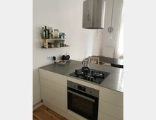 Moderne Küche mit weißem Unterschrank, integriertem Backofen und einem Gasherd mit zwei Kochfeldern auf einer Edelstahl-Arbeitsplatte. Darüber hängt eine silberne Dunstabzugshaube, links befinden sich Wandregale mit Gewürz- und Vorratsgläsern, und im Hintergrund ist ein Esstisch sowie eine Wanduhr sichtbar.