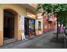 Straßenszene mit einem kleinen Laden mit der Aufschrift "Back Tabak" und weiteren Schildern, die Backwaren und Getränke bewerben. Das Gebäude hat eine gelblich-rote Fassade, und neben dem Eingang befinden sich Verkaufsplakate; links ist ein weiterer Hauseingang mit Holzrahmen sichtbar.