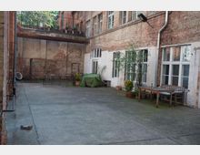 Hinterhof eines Gebäudes mit Ziegelsteinfassaden, großen weißen Fenstern und einer Betonfläche. Im Hof stehen Gartenmöbel, Topfpflanzen und eine Abdeckung über einem Gegenstand; ein Metallgerüst und eine Mauer sind im Hintergrund zu sehen.