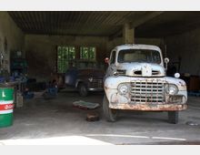 Ein alter, rostiger Lieferwagen und ein klassischer PKW stehen in einer offenen, schlicht gestalteten Werkstatt mit Betondecke. Im Hintergrund befinden sich Arbeitswerkzeug, Regale und große Fenster, die etwas Licht hereinlassen.