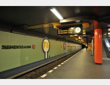 U-Bahnhof Siemensdamm in Berlin mit einer modernen Gestaltung, hellgrünen Wänden mit kreisförmigen Grafiken und leuchtend roten Säulen. Im Vordergrund sind der Bahnsteig, ein Gleis, eine Rolltreppe sowie eine Anzeigetafel und eine Uhr zu sehen.