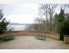 Ziegelstein-Terrasse mit halbkreisförmiger Brüstung, umgeben von Bäumen mit Blick auf einen See im Hintergrund; zwei grüne Metallbänke stehen sich gegenüber auf dem Kiesboden.