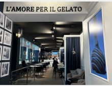 Ein modern eingerichtetes Eiscafé mit dunklem Interieur und eleganten blauen Vorhängen. Im Raum befinden sich Tische und Stühle sowie Sofas auf der rechten Seite; an der Wand hängt der Schriftzug „L’Amore per il Gelato“.