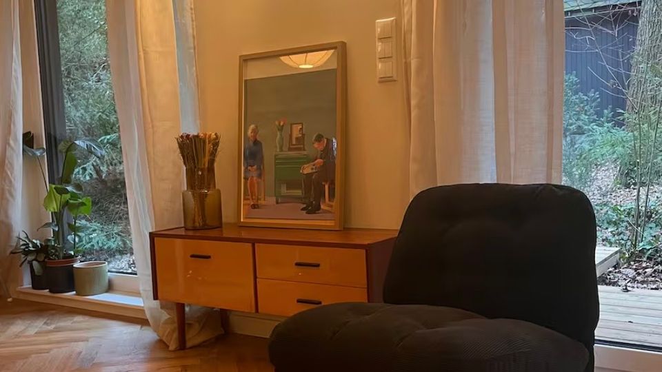 Modernes Wohnzimmer mit Holzparkettboden, einem schwarzen Sessel und einem Holz-Sideboard mit einem Spiegel darauf. Große Fenster mit weißen Vorhängen bieten Blick auf einen bewaldeten Garten.