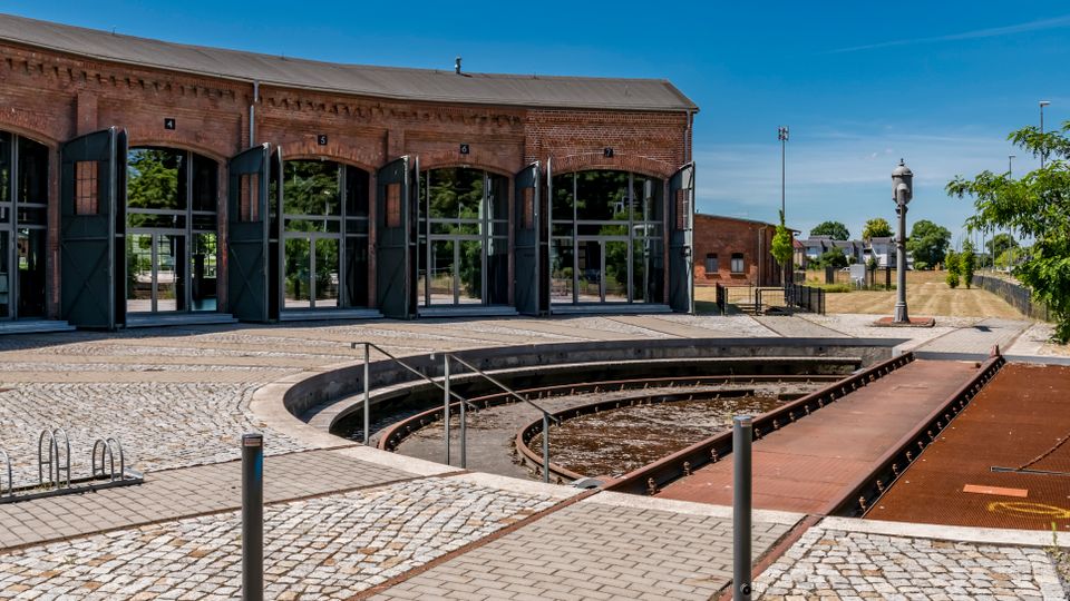 Backstein-Ringlokschuppen mit mehreren hohen Toren und großen Glasflächen, davor eine halbkreisförmige Drehscheibe mit Schienen in einem gepflasterten Bahnhofsareal. Rechts sind ein Gehweg, Laternen, Grünflächen und weitere niedrige Gebäude unter blauem Himmel zu sehen.