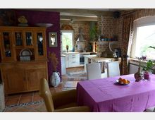 Gemütlicher Wohn- und Küchenbereich mit rustikalem Stil, gekennzeichnet durch freiliegende Backsteinwände, Holzmöbel und lila Akzente. Im Vordergrund ein Esstisch mit pinker Tischdecke, dahinter eine offene Küche mit weißer Einrichtung und Edelstahl-Dunstabzugshaube.