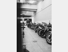 Schwarz-weiß-Foto eines Innenraums mit einer Reihe geparkter Motorräder auf beiden Seiten eines gefliesten Ganges. Die Wände sind schlicht weiß, die Decke zeigt sichtbare Gebrauchsspuren, und im Hintergrund ist ein Bereich mit großen Fenstern und Tischen erkennbar.