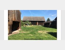 Hof mit großzügiger Rasenfläche, umgeben von mehreren Holzbauten, darunter eine große Scheune mit Ziegeldach im Hintergrund. Rechts und links stehen kleinere, teils offene Holzgebäude, und in der Mitte befinden sich ein Tisch, Stühle und eine Rutsche im Schatten eines blühenden Strauchs.