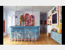 Bunt dekorierter Raum mit einer blauen, marmorierten Bar und transparenten Hockern. Im Hintergrund befinden sich leuchtend rote, bogenförmige Regale mit Spirituosen, daneben Wände mit gerahmten Kunstwerken und Möbeln in Pastellfarben.