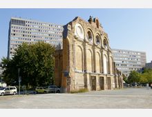 Ruine einer historischen Fassade aus gelbem Backstein mit großen Rundbögen und runden Fenstern, umgeben von modernen Wohngebäuden und Bäumen in einem urbanen Umfeld. Im Vordergrund ist ein Parkplatz mit geparkten Autos und eine gepflasterte Freifläche zu sehen.