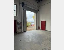 Innenraum einer großen Industriehalle mit Betonboden und weiß verputzten Wänden, dominiert von einem halb geöffneten Rolltor mit Blick nach draußen auf herbstliches Laub und ein Gewässer. Rechts steht eine rote Tür neben einem Feuerlöscher, links ein Fenster und eine auf einer Staffelei aufgestellte Tafel.