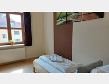 Schlicht eingerichtetes Zimmer mit einem Einzelbett, darüber ein Bild mit Naturmotiv. Ein großes Fenster mit Holzrahmen und Blick auf benachbarte Gebäude, darunter ein Heizkörper und eine kleine Zierpflanze.