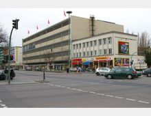 Straßenszene mit einem modernen mehrstöckigen Gebäude mit Fassaden aus beigen Platten und horizontalen Fenstern; im Erdgeschoss befinden sich Geschäfte. Daneben steht ein kleineres weißes Gebäude mit Schaufenstern, über denen Werbeschilder hängen. Autos und Fußgänger sind an einer Kreuzung mit Ampeln sichtbar.