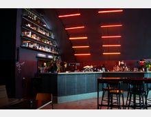 Innenraum einer modernen Bar mit dunkler Farbgestaltung. Eine lange Theke mit verschiedenen Flaschen und Gläsern wird von Regalen an der Wand begleitet, während rote Neonlichter von der Decke hängen und den Raum beleuchten.