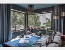 Wohnzimmer mit moderner Einrichtung, großen Eck-Fenstern und Blick auf eine grüne Außenanlage. Der Raum ist mit einem blauen Teppich, einer grauen Couch mit bunten Kissen und schwarzen Sesseln gestaltet; an den Fenstern hängen lange blaue Vorhänge.