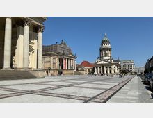 Breiter Platz mit symmetrischem Pflastermuster, umgeben von historischen Gebäuden: links ein Gebäude mit Säuleneingang, in der Mitte ein Opernhaus mit einem Gerüst, und rechts der Französische Dom mit Kuppel. Der Himmel ist klar und blau.