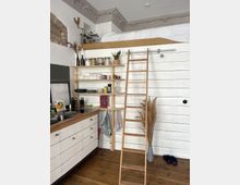 Kleine Wohnung mit offenem Holz- und Weiß-Thema, inklusive einer eingebauten Küche mit weißen Schränken, offenen Regalen mit Geschirr und Vorratsgläsern, einer Leiter zu einem Hochbett und dekorativen Stuckelementen an der Decke. Auf dem Holzfußboden stehen eine Vase mit getrockneten Gräsern und ein Paar Hausschuhe.