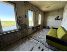 Ein verfallener Raum mit beschädigten Wänden, sichtbaren Betonböden und drei Fenstern, die eine Aussicht auf eine Landschaft mit Wasser und Bäumen bieten. Im Raum stehen ein grünes Sofa und ein schwarzer Kronleuchter an der Decke.