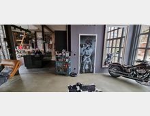 Industrie-inspirierter Wohnraum mit grauer Wand und großen Fenstern, davor ein Motorrad platziert. Auf der Wand befindet sich eine Tür mit einem auffälligen Schwarz-Weiß-Fotodruck, daneben steht ein kleiner blauer Schrank. Im Hintergrund ist eine offene Küche mit sichtbar freiliegenden Holzbalken und Dekorationsgegenständen.