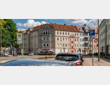 Straßenecke mit mehrstöckigen Wohngebäuden im Altbaustil, roten Ziegeldächern und einem kleinen Geschäft mit gestreifter Markise im Erdgeschoss. Im Vordergrund geparkte Autos, ein Haltestellenschild für den öffentlichen Nahverkehr und ein Parkplatzschild für Menschen mit Behinderung.