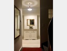 Blick in einen Flur mit hellen Wänden, in dem ein weißes Sideboard mit zwei Türen und einer Schublade steht. Darüber hängt ein goldgerahmter Spiegel, und auf dem Sideboard steht eine Vase mit Blumen. Eine Deckenleuchte beleuchtet den Raum, und ein roter Teppich liegt auf dem Boden.