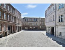 Pflastersteinstraße in einem historischen Stadtensemble, gesäumt von mehrstöckigen Gebäuden mit klassizistischen Fassaden aus Putz und Stein; im Hintergrund ein beige-graues Gebäude mit dekorativem Eingang.