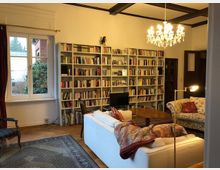 Wohnzimmer mit hellen Wänden und dunklen Deckenbalken, einem Bücherregal, das die gesamte Wand einnimmt, einem weißen Sofa und einem gepolsterten Sessel. Eine Fensterfront mit Vorhängen lässt Licht herein, ein Teppich bedeckt den Holzboden, und ein Kronleuchter hängt von der Decke.