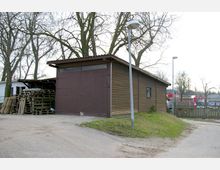 Holzgebäude mit brauner Fassade und großen Doppeltüren, umgeben von kahlen Bäumen. Neben dem Gebäude liegen gestapelte Paletten, und im Hintergrund sind Zäune, Laternen und Gebäudeteile sichtbar.