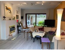 Wohnzimmer mit heller Einrichtung, bestehend aus einer beigefarbenen Eckcouch mit bunten Kissen, einem kleinen Tisch mit Tischdecke, Kamin mit goldener Einfassung und Dekorationen. Große Glas-Schiebetür im Hintergrund mit Blick auf einen Garten.